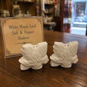 May include: Two white ceramic salt and pepper shakers shaped like maple leaves, each featuring small holes on top. A rectangular sign stands behind them with the printed text: White Maple Leaf Salt & Pepper Shakers, framed by decorative borders.