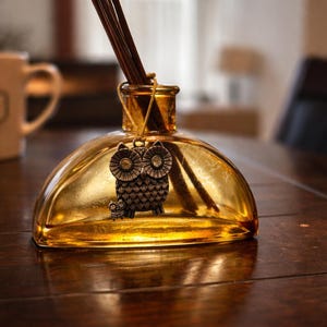 Puede incluir: Un difusor de vidrio ámbar decorativo con un dije de búho de metal. La botella contiene cañas marrones para la difusión de fragancia. El dije de búho está detallado con patrones intrincados. La botella se encuentra sobre una superficie de madera oscura.