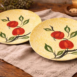 May include: Two round, yellow ceramic plates with a hand-painted design of red fruit and green leaves. The plates are on a tan linen cloth, with a wooden table in the background. The plates are a decorative kitchen item.
