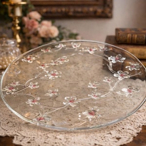 Puede incluir: Una fuente de vidrio transparente con un diseño floral. La fuente presenta delicadas ramas con flores blancas y centros rojos. La marca Mikasa es visible. La fuente está sobre una mesa con un tapete de encaje.