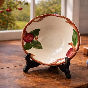 May include: A decorative ceramic bowl with a cream-colored interior and a brown rim. The bowl features hand-painted red apple and green leaf designs. It is displayed on a black stand, showcasing its decorative appeal.