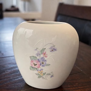 May include: Cream-colored ceramic vase with a rounded shape. The vase features a floral design in pastel pink, purple, and yellow. The flowers are delicately painted on the surface. The vase is sitting on a dark wooden table.