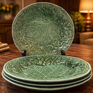 May include: A stack of green ceramic plates with a leaf and grape design. The plates are stacked on a wooden table. One plate is propped up on a black stand. The plates have a detailed, textured surface.