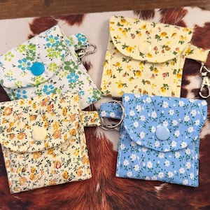 May include: Four small, square fabric pouches with floral patterns in yellow, blue, and white. Each pouch has a button closure and a metal keyring attached. The pouches are arranged on a brown and white patterned surface.