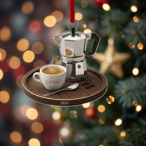 May include: A circular Christmas ornament featuring a silver moka pot, a white coffee cup with the name Jennie printed in gold, a silver spoon, and scattered coffee beans on a dark wood tray. The year 2026 is printed on the edge of the tray.