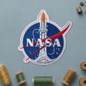 May include: Embroidered NASA patch featuring the NASA logo with a rocket launching into space. The patch is circular with a blue background, white text, and a red swoosh. The rocket is white and orange.