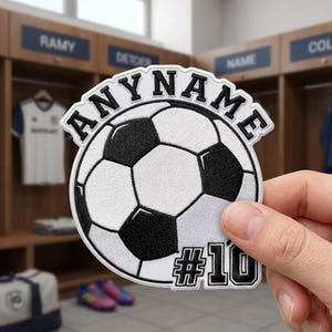 May include: A circular embroidered patch featuring a black and white soccer ball graphic. The text ANYNAME is arched across the top in black block letters with a white border, and the text #10 is positioned at the bottom right in black.