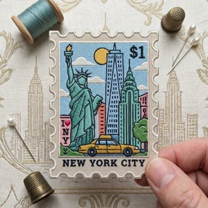 May include: A rectangular embroidered patch shaped like a postage stamp featuring the Statue of Liberty, skyscrapers, a yellow taxi, and the text I NY and NEW YORK CITY with a $1 symbol. The patch is surrounded by a spool of thread and two thimbles.