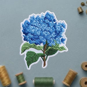 May include: Embroidered patch featuring a vibrant blue hydrangea flower with green leaves and a brown stem. The patch has a white border. Sewing notions, including spools of thread and thimbles, are scattered around the patch.