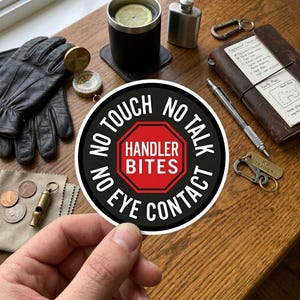 May include: A round black and red sticker with the text "NO TOUCH, NO TALK, NO EYE CONTACT, HANDLER BITES". The sticker is held by a hand. Other items include a leather notebook, compass, gloves, and a flask.