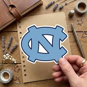 May include: A light blue sticker featuring the interlocking "NC" logo of the University of North Carolina, with a white interior and a dark blue outline. The sticker is held by a hand on a wooden surface.