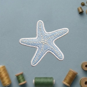 May include: A light blue starfish patch featuring intricate white embroidered details and a textured surface. The starfish has five arms and is surrounded by several wooden spools of thread in various shades of green and gold.