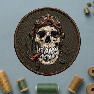 May include: Embroidered patch featuring a skull wearing a vintage aviator helmet and goggles, with a lit cigar in its mouth. The text "TALLY HO" is embroidered on the teeth. The patch is circular with a brown border and a green background.