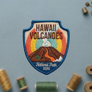 May include: A shield-shaped embroidered patch featuring a brown volcano with orange lava, a green palm tree, and a rainbow background. The text reads HAWAII VOLCANOES NATIONAL PARK 1916. The patch has a blue border and is surrounded by sewing thread spools.