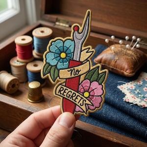 May include: An embroidered patch featuring a red seam ripper, a blue flower, and a pink flower, with a beige banner across the center displaying the text No REGRETS in black lettering. The patch has a gold-colored border and detailed stitching.