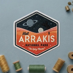 May include: Embroidered patch with a hexagonal shape featuring a night sky with a planet and a figure on a desert landscape. The text reads "Visit Arrakis National Park" and "The Spicy Planet!" in white and orange thread.