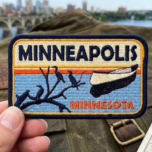 May include: A rectangular embroidered patch featuring the text MINNEAPOLIS in dark blue letters at the top and MINNESOTA in orange letters at the bottom. The design depicts a black canoe and three black birds perched on a dark blue tree branch.