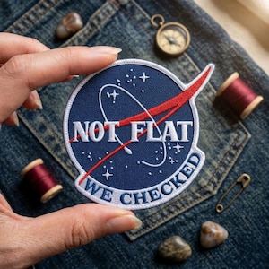 May include: A circular embroidered patch with a dark blue background, featuring a white border and the text NOT FLAT WE CHECKED in white. A red swoosh crosses the center, accompanied by white star and orbital line graphics on the fabric surface.