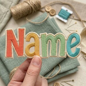 May include: A custom embroidered patch featuring the word Name in a colorful, multi-toned design. The letters are stitched in orange, yellow, light green, and teal, each outlined with a thick, cream-colored border on a fabric base.