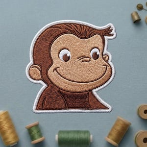 May include: Embroidered patch featuring a smiling monkey with brown fur and a white border. The monkey has large, expressive eyes and a cheerful expression. The patch is likely for clothing or accessories.