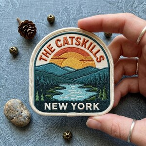 May include: An embroidered patch featuring a landscape scene of The Catskills, New York. The design includes a yellow and orange sun rising over dark teal mountains and a blue lake surrounded by pine trees, with the text THE CATSKILLS NEW YORK.