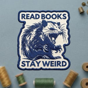 May include: Embroidered patch featuring a blue opossum with an open mouth, surrounded by the text "READ BOOKS STAY WEIRD" in a white and blue design. The patch is on a light blue background.
