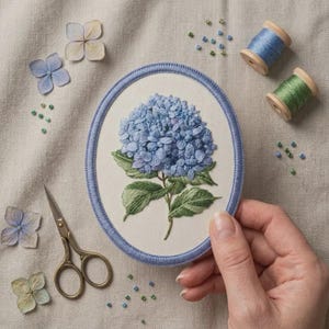 May include: An oval embroidery hoop containing a fabric panel with a detailed blue hydrangea flower and green leaves. The hoop has a blue border. Nearby are two wooden spools of blue and green thread, small scissors, and loose beads.