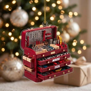 May include: A red tool chest ornament featuring an open lid with various tools, multiple drawers, and a gold hanging string. The chest displays the text NAME and 2026 in white lettering on the front drawers, showcasing a detailed miniature design.