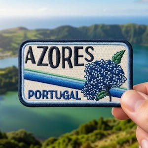 May include: A rectangular embroidered patch with a dark blue border, featuring the text AZORES and PORTUGAL in black lettering. The design includes a cluster of blue hydrangea flowers and a diagonal blue and green stripe on a cream background.