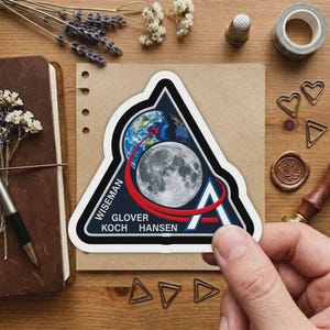 May include: A triangular sticker featuring a design of Earth, the Moon, and a red arc. The sticker has the names WISEMAN, GLOVER, KOCH, and HANSEN. The background includes a notebook, dried flowers, and stationery.