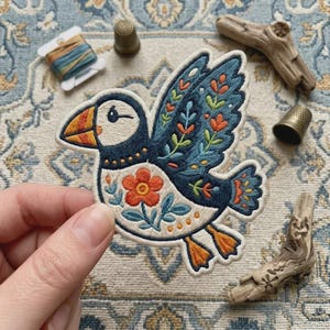May include: An embroidered puffin patch featuring a cream-colored body with a dark blue wing and tail. The design includes orange and green floral motifs, an orange beak, and orange feet, all outlined in white thread on a fabric backing.
