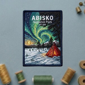 May include: A rectangular embroidered patch featuring a nighttime landscape of Abisko National Park. The design displays a vibrant green aurora borealis, a yellow moon, snow-covered ground, pine trees, and a red tent with the text ABISKO National Park EST. 1909.