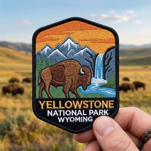 May include: A hexagonal embroidered patch featuring a brown bison standing in front of a blue waterfall, snow-capped mountains, and an orange sky. The black border contains the yellow and white text: YELLOWSTONE NATIONAL PARK WYOMING.