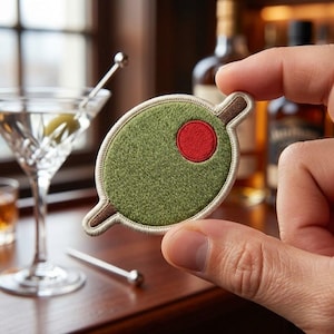 May include: An embroidered patch shaped like a green olive with a red pimento center, featuring a brown toothpick running through the middle. The patch has a cream-colored stitched border and a textured fabric surface with detailed thread work.