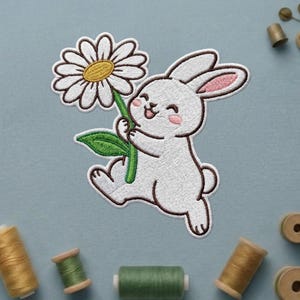 May include: Embroidered patch featuring a white bunny holding a large daisy. The bunny has pink inner ears and rosy cheeks. The daisy has white petals and a yellow center. The patch is on a light blue background with spools of thread.