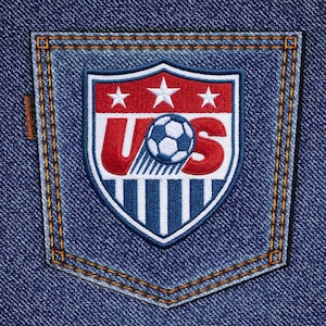 May include: A blue denim pocket featuring an embroidered United States Soccer Federation crest. The shield-shaped patch displays the letters US in red, a white soccer ball, three white stars, and vertical blue and white stripes on a red background.