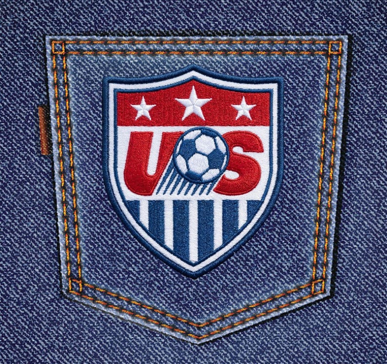 May include: A blue denim pocket featuring an embroidered United States Soccer Federation crest. The shield-shaped patch displays the letters US in red, a white soccer ball, three white stars, and vertical blue and white stripes on a red background.
