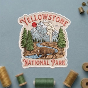 May include: Embroidered patch featuring a Yellowstone National Park design. The patch includes a bison drinking from a river, mountains, trees, and the text "Yellowstone National Park" in a vintage style.