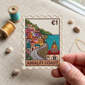 May include: A rectangular embroidered patch shaped like a postage stamp featuring a colorful Amalfi Coast scene with stacked buildings, a winding road, a domed church, and the text €1 and AMALFI COAST in brown thread on a cream background.