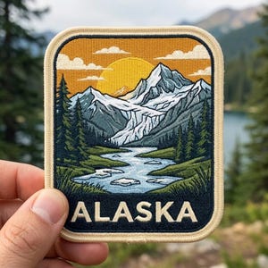 May include: A rectangular embroidered patch featuring a landscape of snow-capped mountains, a winding river, and evergreen trees under a yellow sun and orange sky. The word ALASKA is stitched in large, cream-colored capital letters at the bottom.