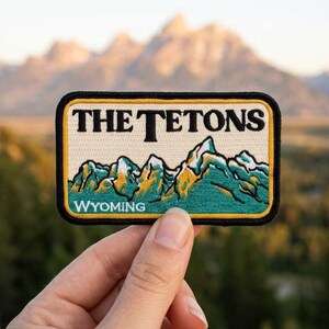 May include: A rectangular embroidered patch featuring a cream-colored background with the black text THE TETONS at the top and WYOMING at the bottom. The center displays a stylized mountain range in shades of teal, green, and gold with white peaks.