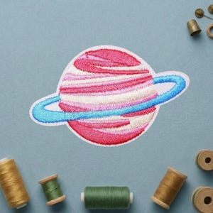 May include: Embroidered patch featuring a planet design with pink, white, and red stripes, a blue ring, and a white border. The patch is on a light blue background, with spools of thread and sewing notions scattered around.