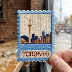 May include: A rectangular embroidered patch shaped like a postage stamp with a light blue border. The design features the Toronto skyline in shades of gold, brown, and dark blue against a cream background, with the word TORONTO in brown text.