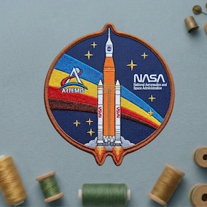 May include: Embroidered patch featuring the NASA Artemis program logo. The patch is circular with a blue background, a rocket, and the NASA logo. The design includes orange, yellow, and white colors, with the word "ARTEMIS" in white.