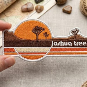 May include: An embroidered patch featuring a desert landscape with a large orange sun, brown silhouettes of Joshua trees, and horizontal stripes in shades of orange, cream, and brown. The text joshua tree is stitched in white on the right side.