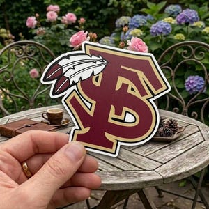May include: A maroon and gold sticker featuring the letters "FS" with a feather design. The sticker is held in front of a blurred background of a garden with flowers and a wooden table.