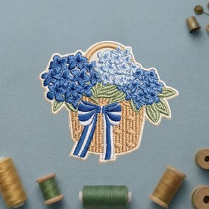 May include: An embroidered patch featuring a tan woven basket filled with clusters of light and dark blue hydrangea flowers, green leaves, and a prominent blue and white striped ribbon tied in a bow at the front of the basket.