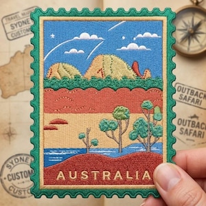 May include: A rectangular embroidered patch shaped like a postage stamp with a green scalloped border. The design features a blue sky with clouds, tan rock formations, green trees, and red desert terrain. The word AUSTRALIA is stitched in gold.