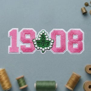 May include: Pink embroidered patch with the numbers "1908" and a green and white emblem. The patch is surrounded by spools of thread in various colors, including gold and green.