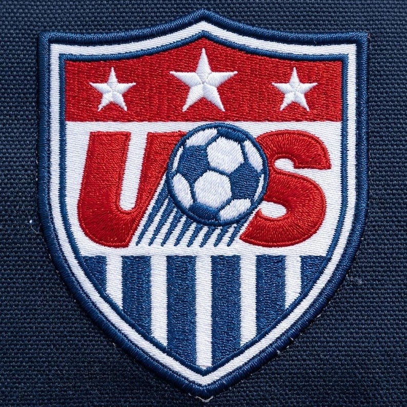 May include: An embroidered shield-shaped patch featuring the letters US in red with a soccer ball replacing the letter S. The top section is red with three white stars, and the bottom section features alternating vertical blue and white stripes.